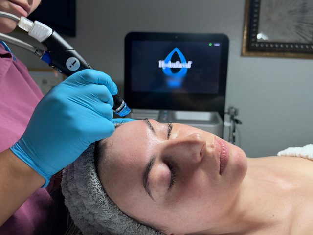 An esthetician in blue gloves moving a HydraFacial wand along a client’s forehead at The Med Spa Wimberley, the HydraFacial machine behind